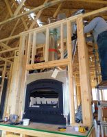 installation photo (2)-min A man standing on a ladder, working on the construction of a wooden room with a fireplace, illustrating a construction or renovation project